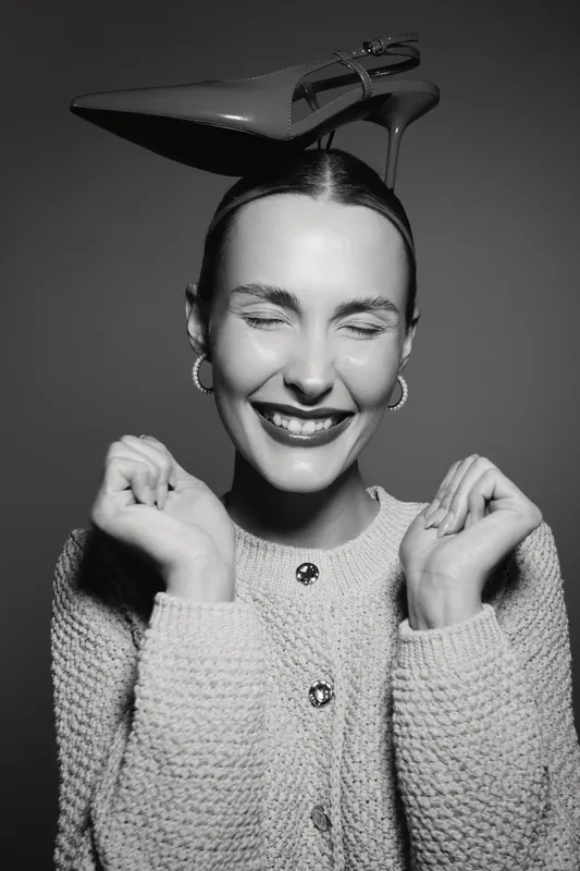 Happy white woman with pale skin and closed eyes smiling wearing knitted sweater and high heel shoe on her head in black and white studio photo.