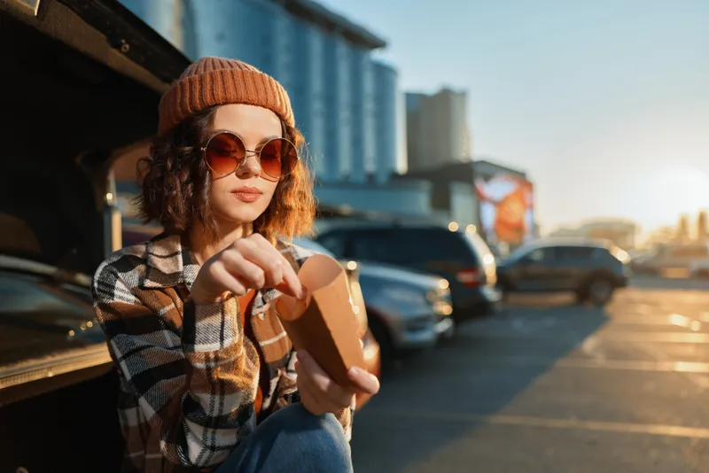 Woman with sunglasses and beanie at car trunk in parking lot eating a snack during golden hour glow, candid lifestyle portrait with authenticity, mindful living and emotional storytelling mood.