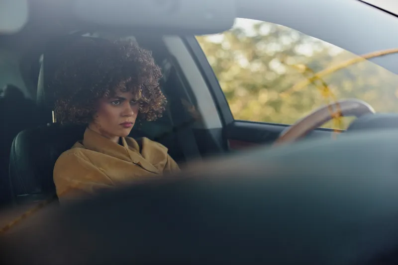Woman sits inside a car with retro film color palette, calm lifestyle mood, focused gaze, soft light through glass, vintage automotive scene, candid contemplative moment