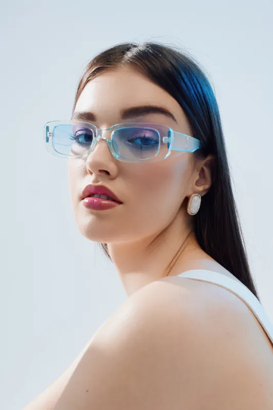 Young woman with long dark hair wears transparent blue glasses and white earrings. Close-up portrait on a light background. Concept of beauty fashion and modern style.