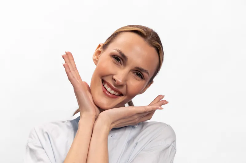 Smiling woman with clean skin poses with hands near face on white background. Beauty portrait with natural makeup and positive expression. Concept of skincare and happiness.