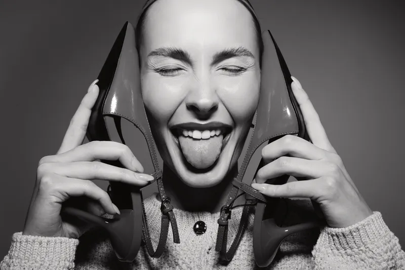 Happy Caucasian woman with light skin holding two shoes near her face and sticking out her tongue. Concept of fun, joy, and playful attitude in monochrome portrait.