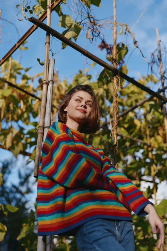 Woman portrait in a colorful sweater outdoors leaning on a vine trellis, casual autumn fashion with relaxed pose, natural sunlight on face and blue sky background.