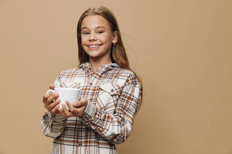 Girl with long hair and plaid shirt smiling and holding white cup with coffee or tea at beige background. Youth, happiness and warm drink concept.