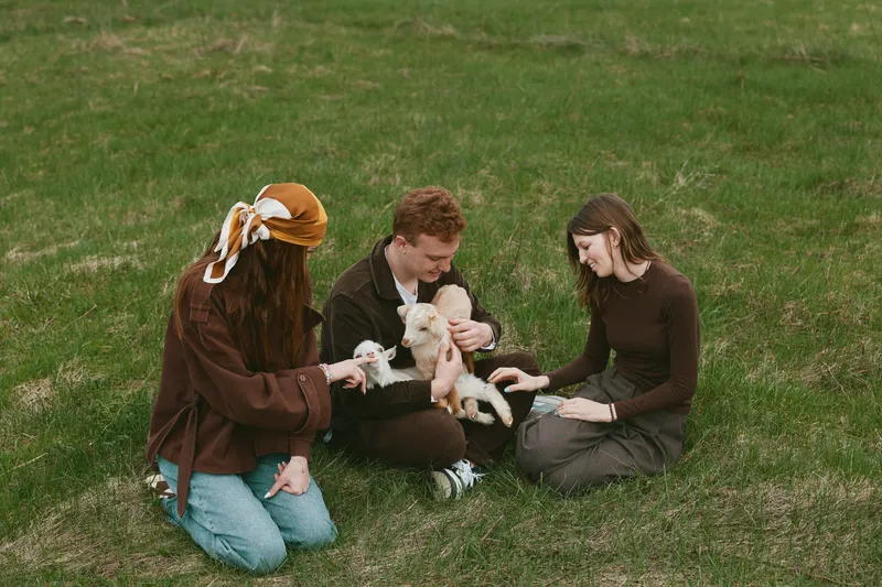 A small goat kid sits with a family in a sunlit park, three people gently petting the animal, creating a warm, casual lifestyle moment outdoors.