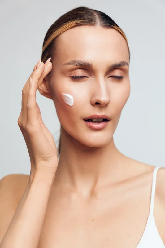 Young woman with clear skin applies moisturizing cream on her face with closed eyes, demonstrating skincare routine and natural beauty on plain light background.