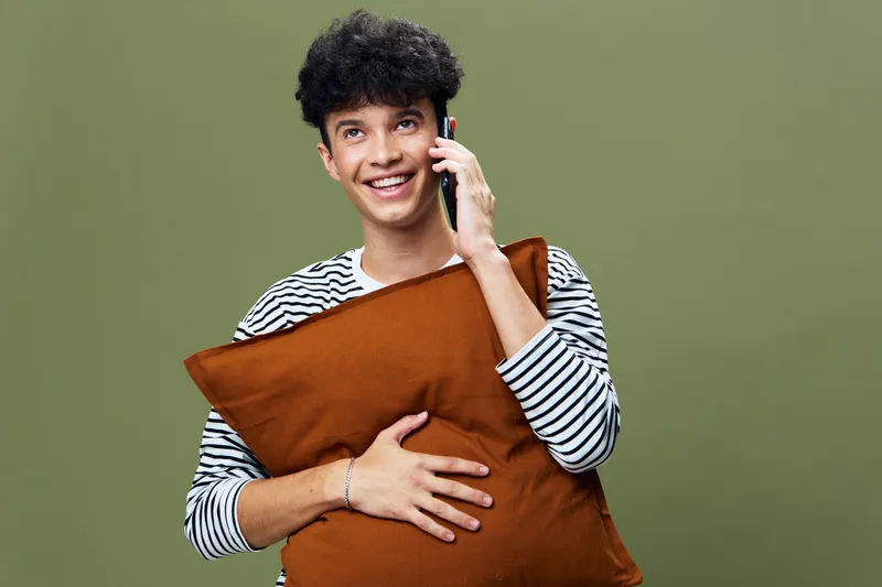 smiling young man talking on smartphone while holding brown pillow against green background in casual striped shirt with joyful expression