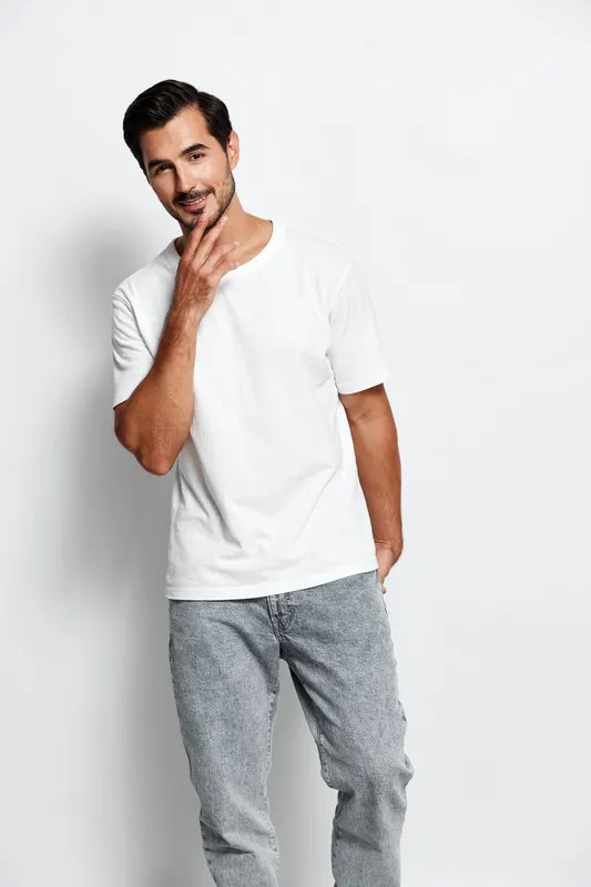 Charming man in casual white t-shirt poses confidently with hand on chin, looking at camera against a minimalistic background. Fashion and lifestyle concept.