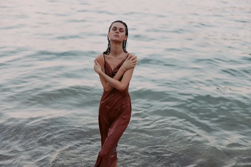 A solitary woman stands waist?deep in calm sea water, wearing a rust brown dress, hair slicked back, embodying serenity, poise, and graceful isolation against a blue horizon.
