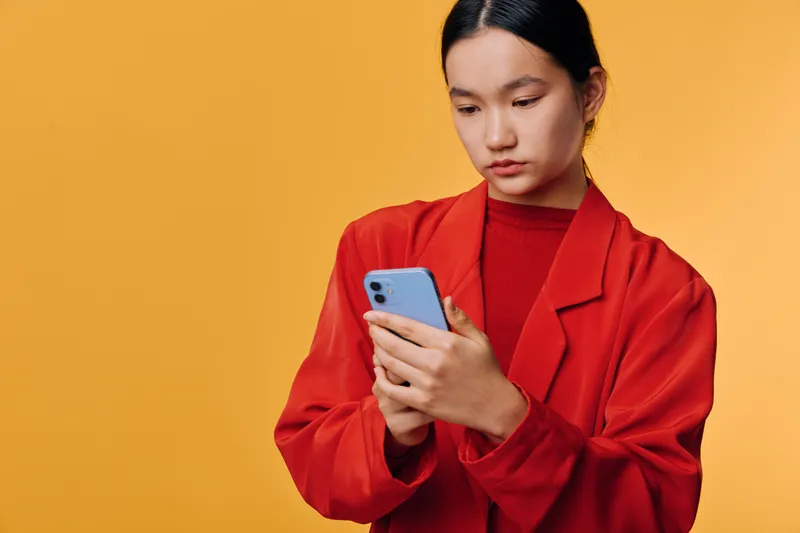 Young woman in red jacket using smartphone on orange background for communication and social media. Female holding mobile phone, focused on screen for technology and connection.