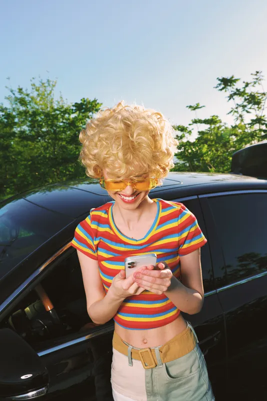 Curly blond girl in striped shirt stands beside a car, looking at a smartphone. Bright outdoor light creates a casual lifestyle mood, playful moment, and modern tech use.
