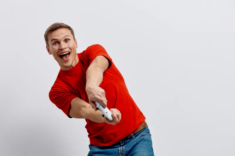 Cheerful young man with short hair, wearing red t-shirt, excited and smiling while holding a TV remote control, expressing fun and happiness isolated on light gray background. Youth lifestyle concept