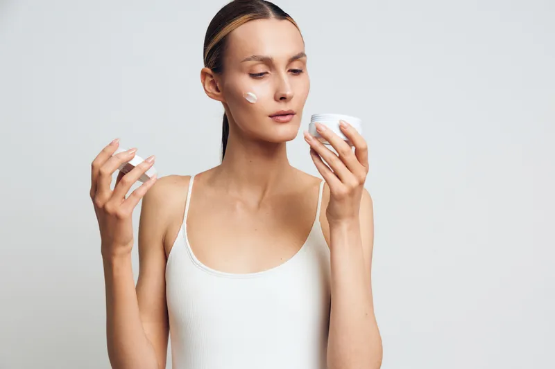 A young woman in a white tank top applies cream on her face while holding a jar of moisturizer. The scene shows skincare routine and self-care with a clean white background.