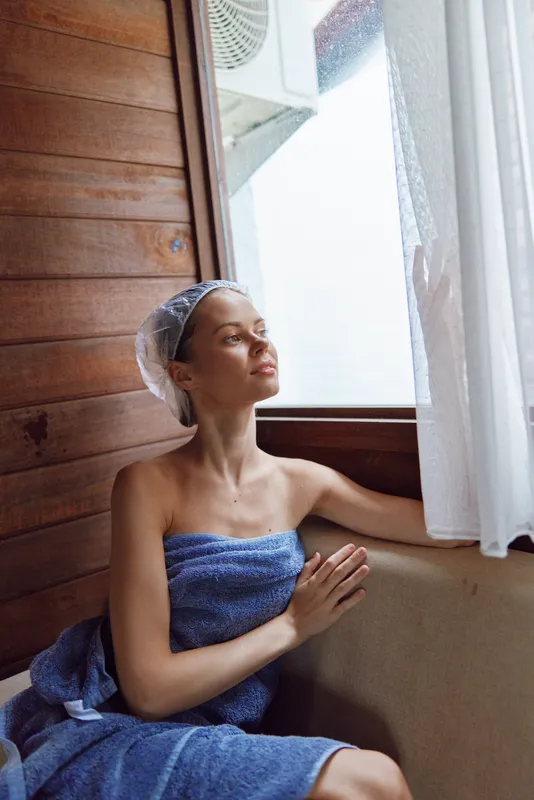 Relaxed woman in a bath towel and shower cap sitting by window in wooden sauna interior, enjoying sunlight and peaceful moment with soft natural light around.
