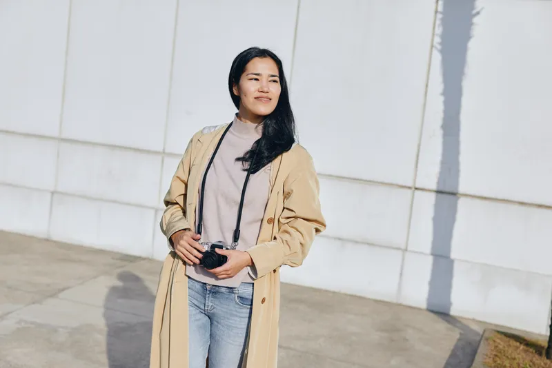Woman standing outdoors with camera in hand, wearing a beige long coat over knit sweater and jeans, sunlit street setting, casual fashion photography moment in daily life.