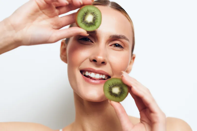 Young woman with blonde hair smiles joyfully while holding two halves of a kiwi fruit near her face against a white background in a bright studio setting.