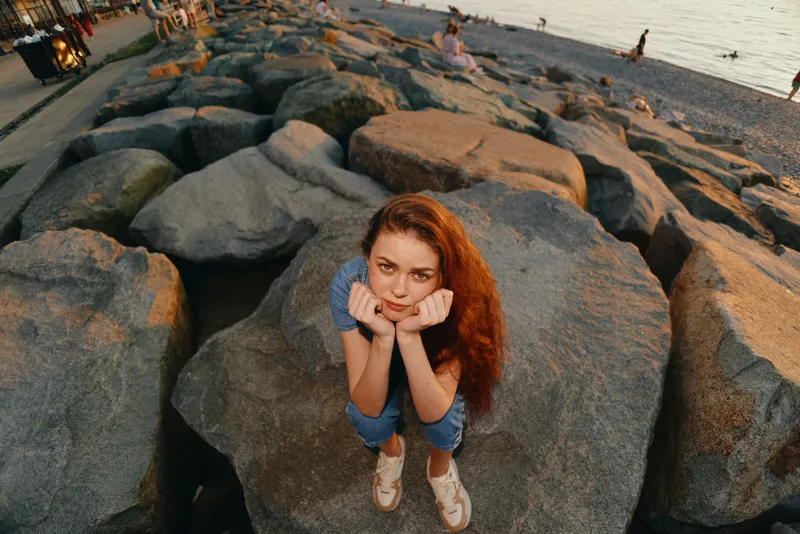 Lifestyle woman in modern boho-western denim street style sitting thoughtfully on large rocks by the beach, warm film color tones highlighting casual summer fashion.