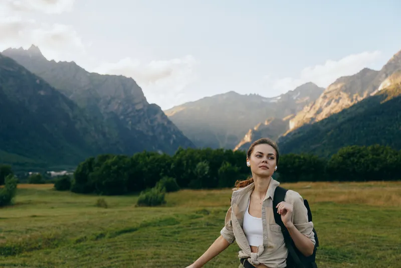 Young woman with backpack hiking in green mountain valley during daytime. Clear sky and sunlight highlight natural landscape and outdoor lifestyle concept.