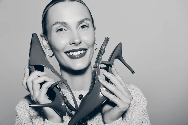 Smiling caucasian woman with light skin holding two high heels near face, wearing sweater and earrings, studio portrait with joyful mood and fashion concept.