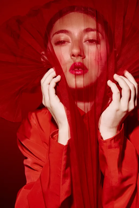 Fashion model in a vibrant red outfit posed behind delicate fabric creating dramatic shadows. Glossy lips and intense gaze convey glamorous beauty and creative style concept.