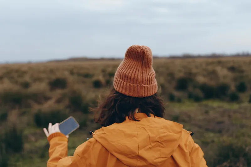Woman outdoor lifestyle, back view, wearing orange jacket and knit beanie, holding smartphone, taking photo of landscape, rolling hills, soft daylight, serene travel mood