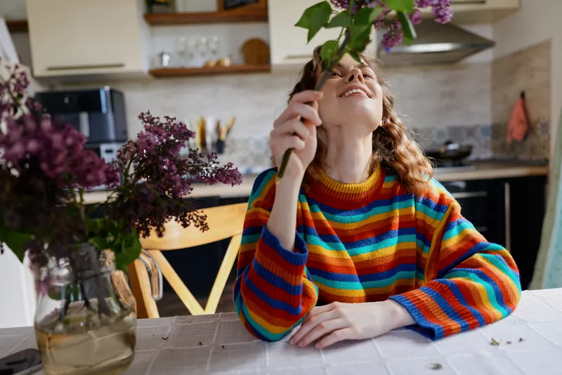 Woman at home admires a flower bouquet, wearing a colorful rainbow striped sweater, a warm kitchen behind, calm lifestyle moment, candid indoor portrait