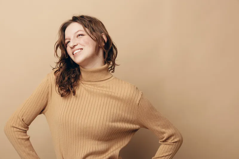 Smiling woman with curly hair wearing beige turtleneck sweater poses confidently against beige background. Concept of happiness, casual style, and natural beauty in studio portrait.