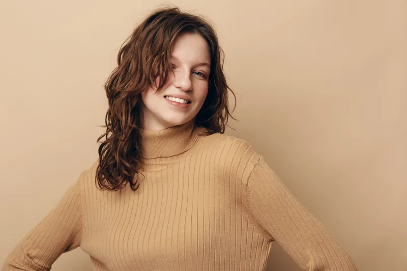 Smiling young woman with curly brown hair wearing beige turtleneck sweater stands against beige background. Casual style, natural beauty, confident and relaxed pose.