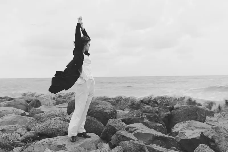 Woman stands on rocky coastline with arms raised, wind blowing her coat behind, capturing freedom, resilience and triumph in an outdoor fashion moment against a vast sea backdrop