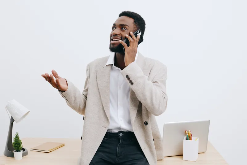 Smiling man in beige suit talking on mobile phone in modern office space with white background, work desk, computer, and potted plant, professional business lifestyle concept