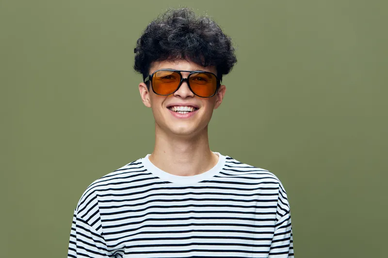 smiling man in striped shirt wearing orange sunglasses with curly hair, happy expression on green background, casual style portrait, youthful and bright atmosphere