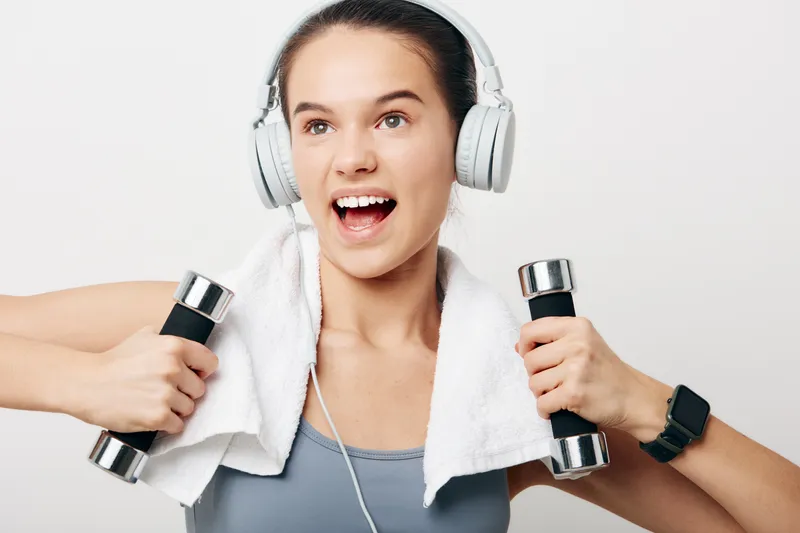 Energetic woman fitness portrait with headphones and towel, holding chrome dumbbells and wearing a smartwatch and sports top, joyful expression on a clean white studio background.