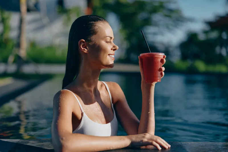 Smiling woman in white swimsuit relaxing in pool holding a red smoothie glass. Sunny outdoor setting with blurred green trees in background. Concept of lifestyle, wellness, and summer relaxation.