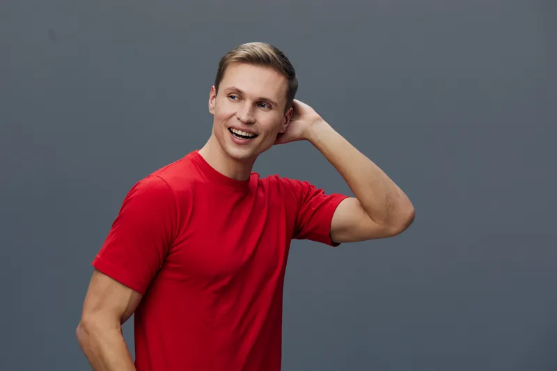 Smiling man wearing red t-shirt standing against gray background touch head happy cheerful casual lifestyle concept mental health and gadgets