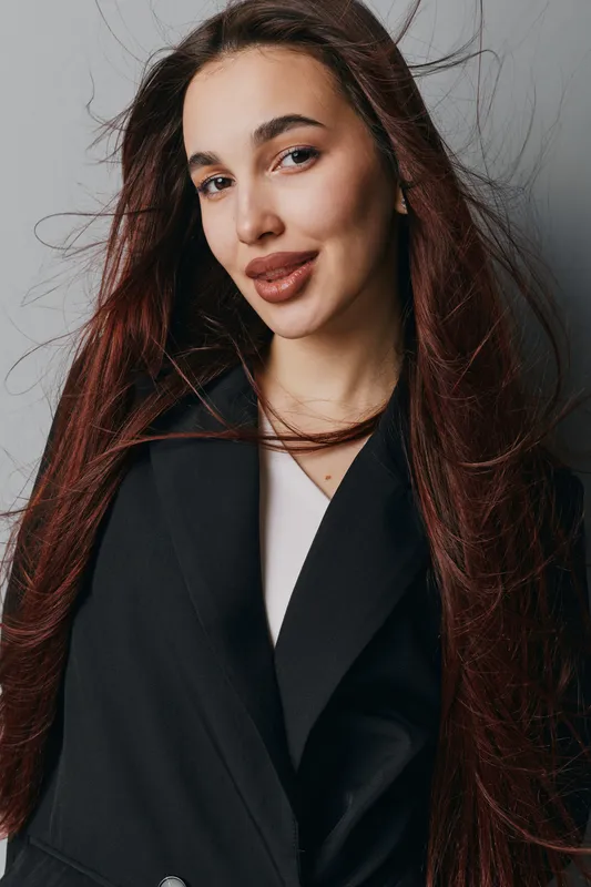 Woman with long brown hair and natural makeup wearing black coat posing indoors against gray wall. Confident female model with casual hairstyle and soft smile for portrait or fashion.