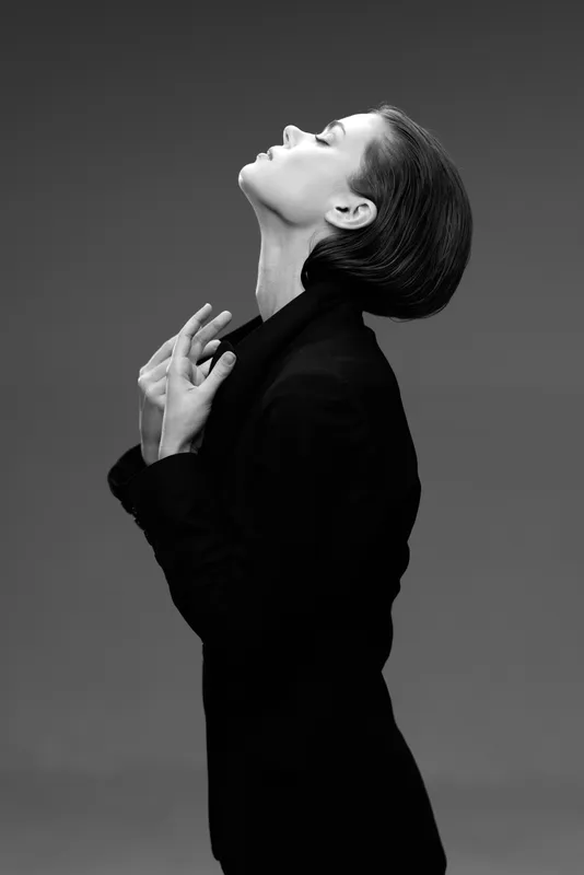 Black and white portrait of a woman in a sleek black outfit posing with eyes closed and head tilted back, expressing elegance and grace.
