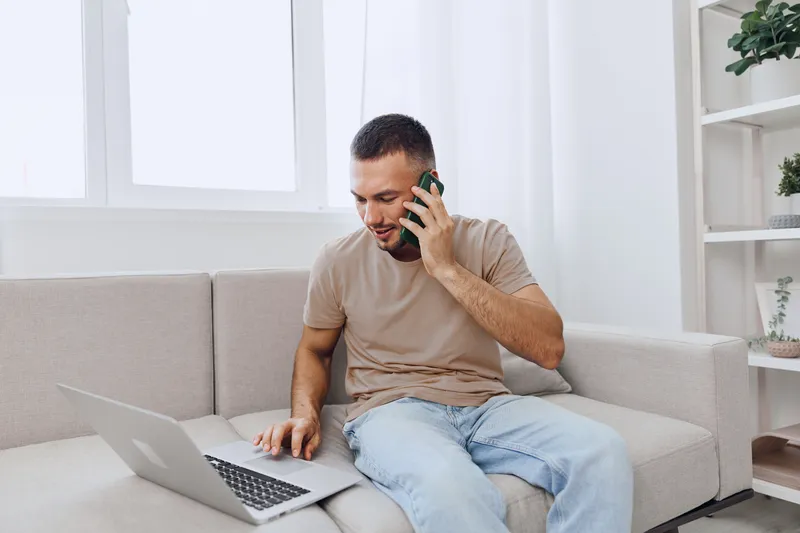 Smiling man talking on the phone while working on a laptop, showcasing modern home office productivity and casual work-life balance in a cozy environment.