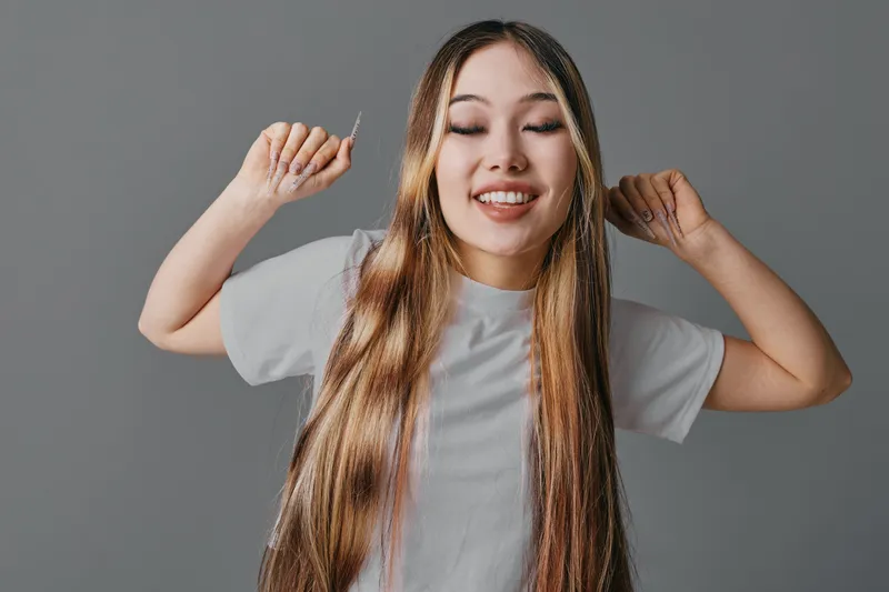 Young woman with long hair and closed eyes smiling while holding hair strands, wearing white t shirt against plain gray background in studio lighting.
