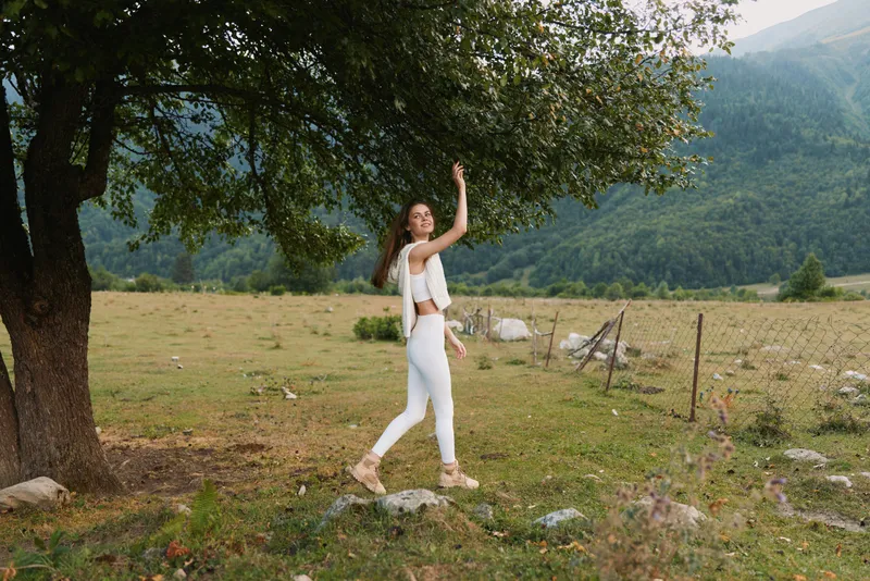 Young woman in white outfit walking under a tree in a green field with mountains in the background. Lifestyle concept with nature, freedom, and outdoor activity.