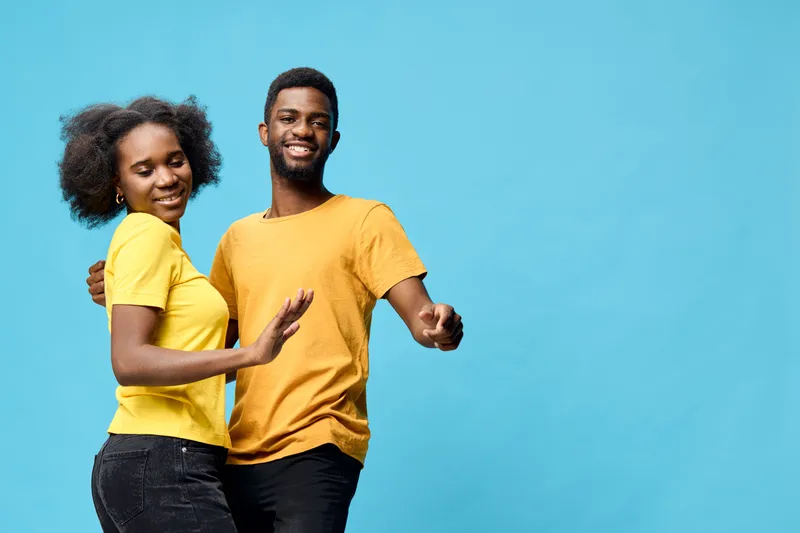 Happy couple dancing together against a vibrant blue background, wearing matching yellow t shirts, showcasing joy and connection in a playful moment