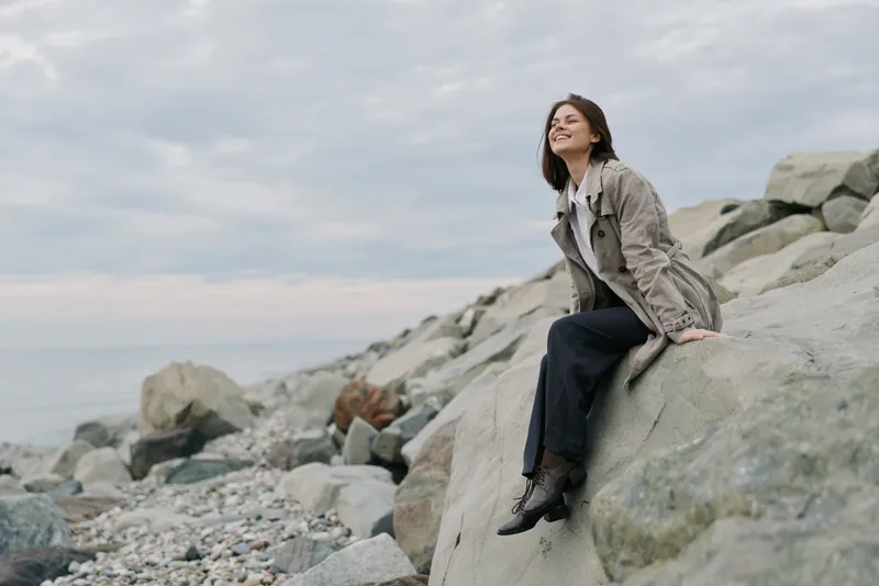 Young woman in casual clothes sitting on a large rock at the rocky beach, smiling and looking away. Overcast sky and calm sea in the background create a peaceful mood.