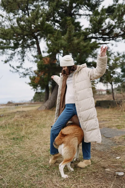 Woman in a long winter coat plays with a dog in an open park, autumn grass and trees, a joyful outdoor moment in nature