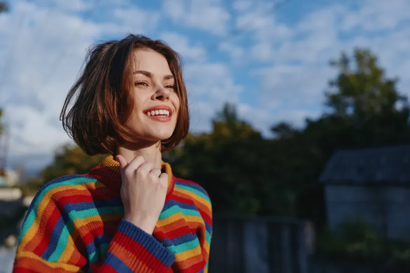 Young woman mid-20s smiling woman in a colorful striped sweater and casual outfit, outdoor portrait with sunlit face, looking up and holding collar, joyful lifestyle and fashion vibe.