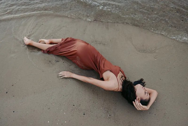 A woman in a rust dress lies on wet sand near the shoreline, relaxed pose, fabric pooling and catching light, creating a calm, minimalist beach portrait.
