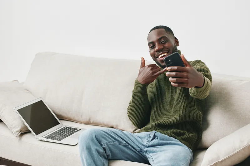 Young Black man in a cozy green sweater taking a selfie at home, exuding happiness and confidence, with a laptop nearby