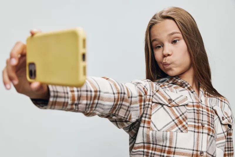 Girl with long hair wearing plaid shirt making selfie with yellow phone, posing with playful facial expression on white background in studio. Child, teen and digital device closeup.