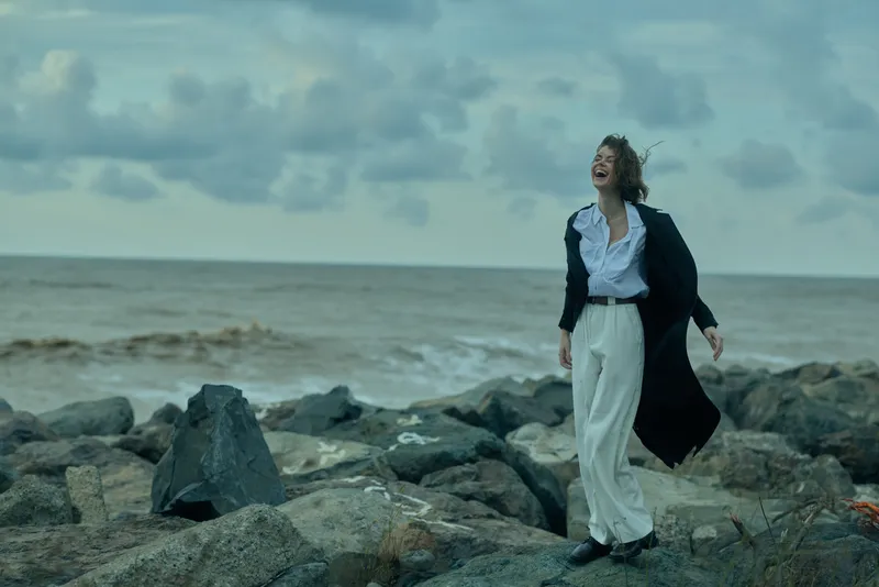 Woman on a rocky shoreline wearing a flowing coat, white pants and a crisp shirt, by the sea in windy weather, conveying fashion and outdoor mood with movement and drama