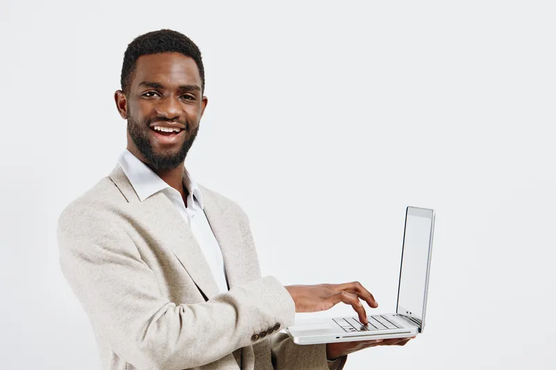 Smiling young man with dark skin using a silver laptop computer on a plain white background. Casual professional look, enthusiastic expression, holding laptop, studio portrait. People lifestyle concep