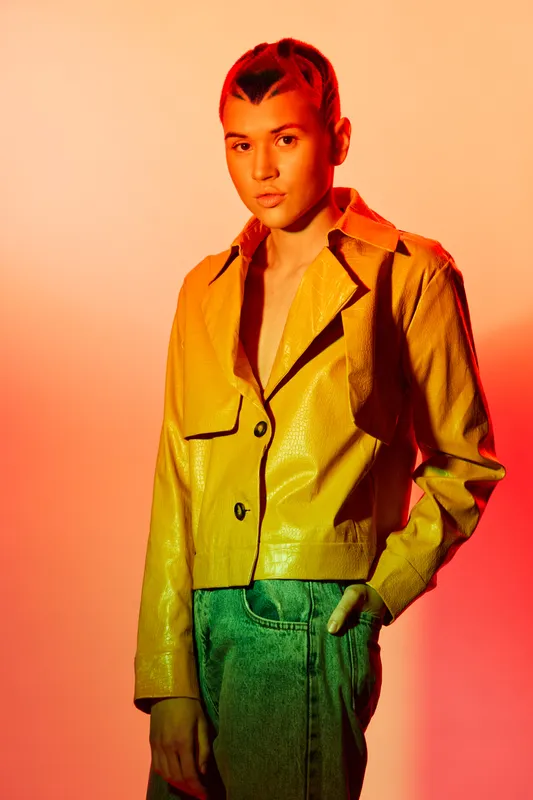Young woman wearing a shiny yellow jacket and green pants stands against vibrant orange background, showcasing modern style and confident posture in studio lighting.