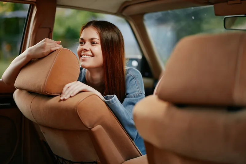 Joyful young woman with long brown hair smiling in a vintage car, wearing denim, conveying happiness and nostalgia in a warm, colorful setting.
